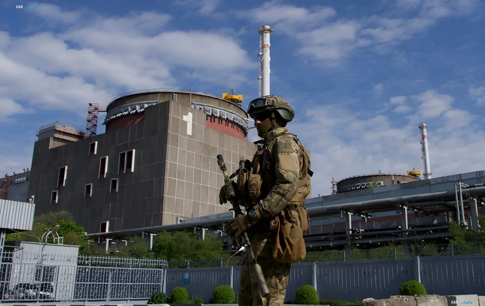Военный в камуфляже стоит на фоне здания Запорожской АЭС, символизируя напряженную обстановку вокруг объекта